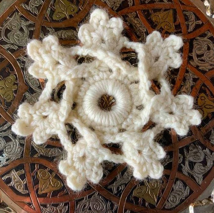 Cremefarbene gehäkelte Schneeflocken-Applikation auf ornamentaler Metalloberfläche.
