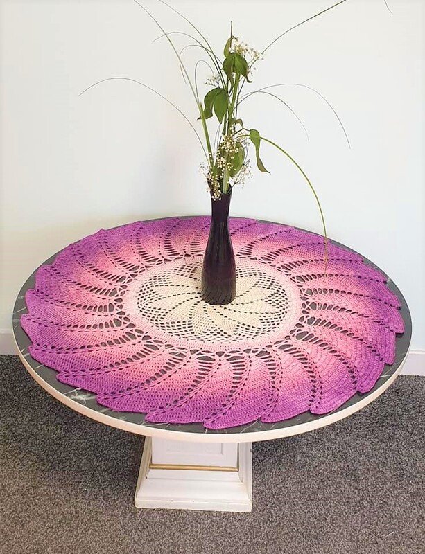 Rundes gehäkeltes Deckchen in Lila, Rosa und Creme mit Spitzenmuster und einer Vase in der Mitte auf einem runden Tisch.