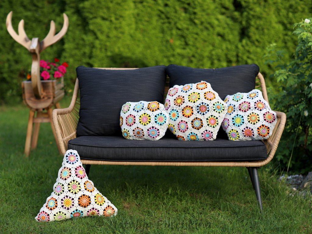 Drei bunte gehäkelte Granny‑Square‑Kissen auf einer dunkelgrauen Gartenbank; ein dreieckiges Kissen liegt im Vordergrund auf dem Rasen