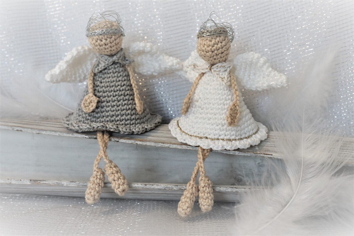 Zwei gehäkelte Amigurumi-Engelchen, eines grau und eines weiß, mit Flügeln und silbernem Kranz, sitzend auf einem Holzbrett
