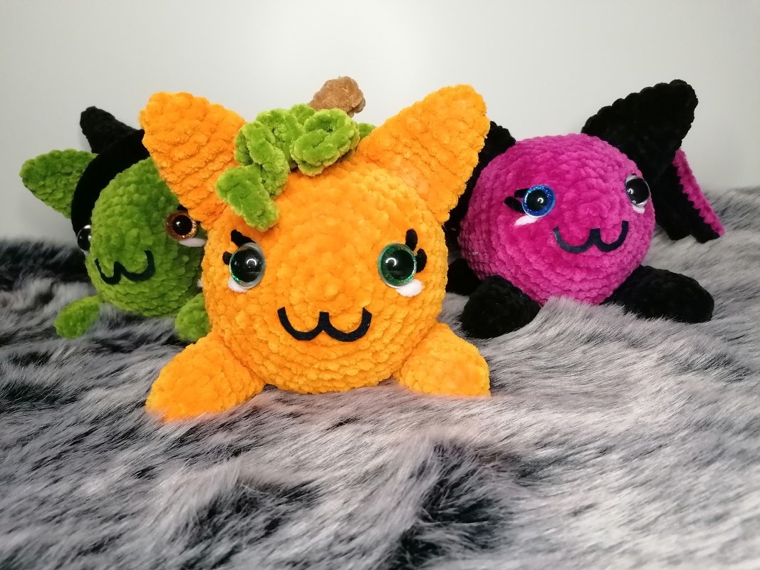Drei runde, flauschige, handgehäkelte Amigurumi-Kürbis-Katzen in Orange, Grün und Pink auf grauem Kunstfell.