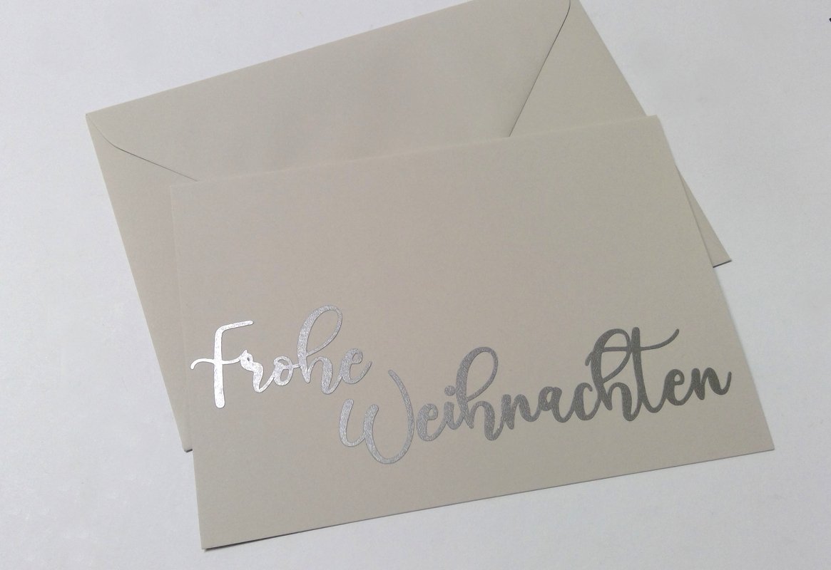 Beigefarbene Karte mit silbernem Schriftzug „Frohe Weihnachten“ und passendem Umschlag