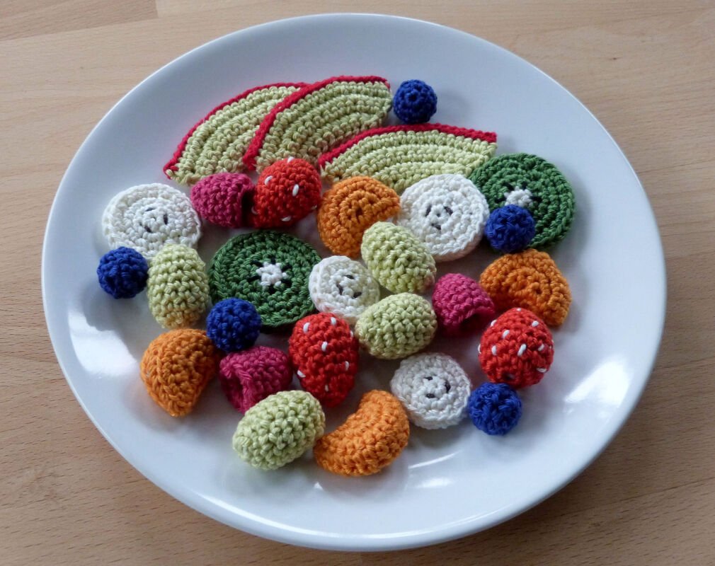 Weißer Teller mit bunten, handgehäkelten Früchten und Gemüsestücken wie Erdbeeren, Kiwischeiben und kleinen Beeren.