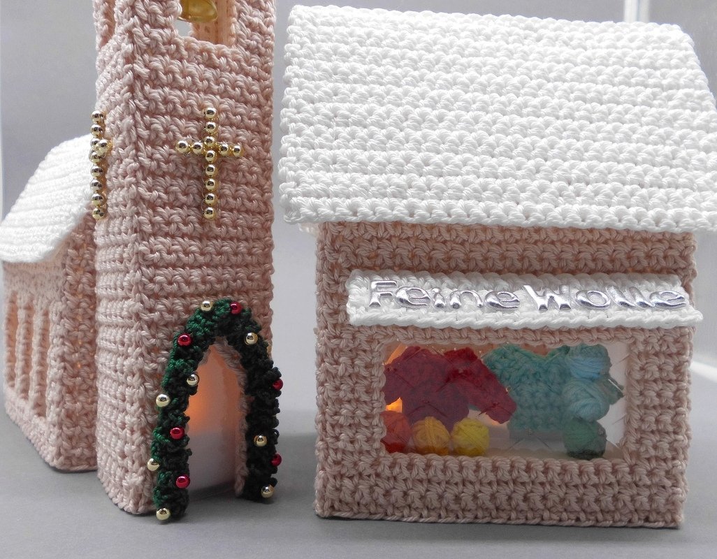 Gehäkeltes Set aus einer rosa Kirche mit Perlenkreuz und einem Haus mit aufgesetztem Schriftzug, beide mit weißen Dächern und Innenbeleuchtung