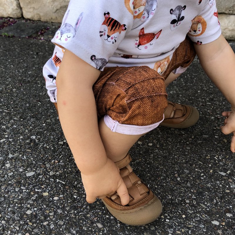 Nahaufnahme eines hockenden Kleinkinds, Fokus auf braune Baby-Shorts mit Umschlag und sichtbarer Naht; Hände greifen zur Sandale.