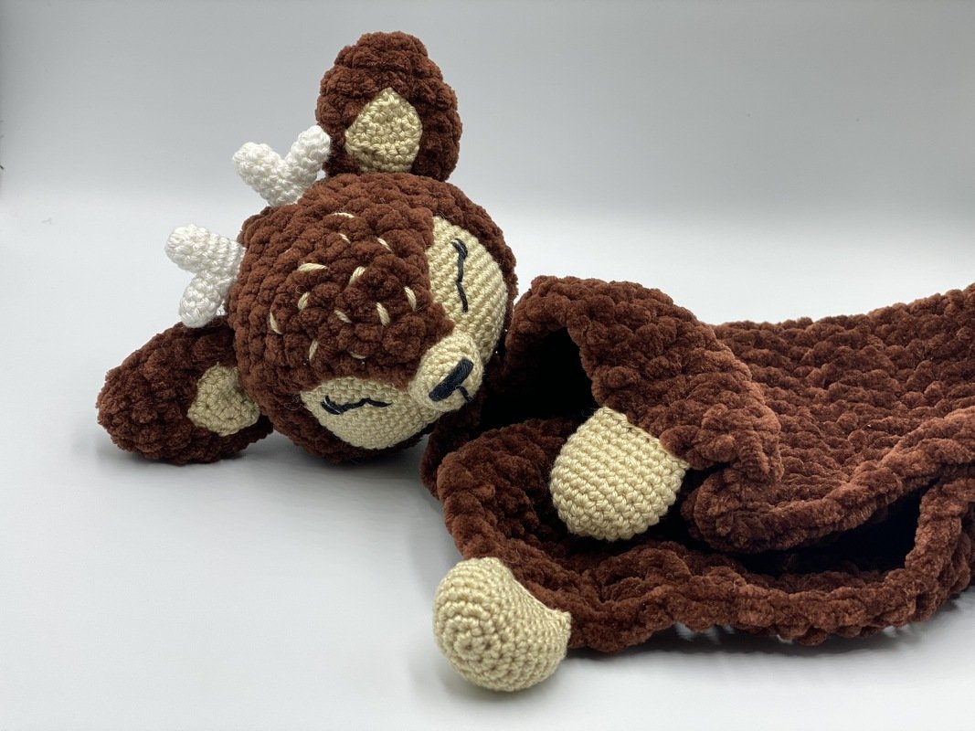 Brown crochet deer snuggle blanket with small white antlers and beige face details lying on a white surface.