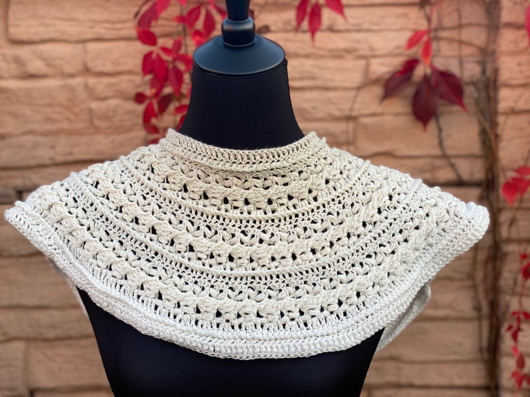 Detailaufnahme eines cremefarbenen, gehäkelten Capelets mit Stehkragen auf einer schwarzen Puppe.