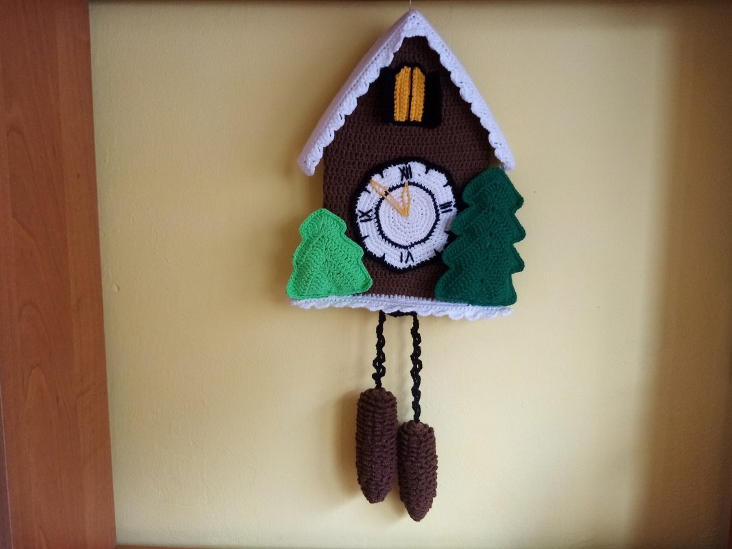 Crocheted cuckoo clock wall hanging with white clock face, green trees, and hanging brown weights on a yellow wall.
