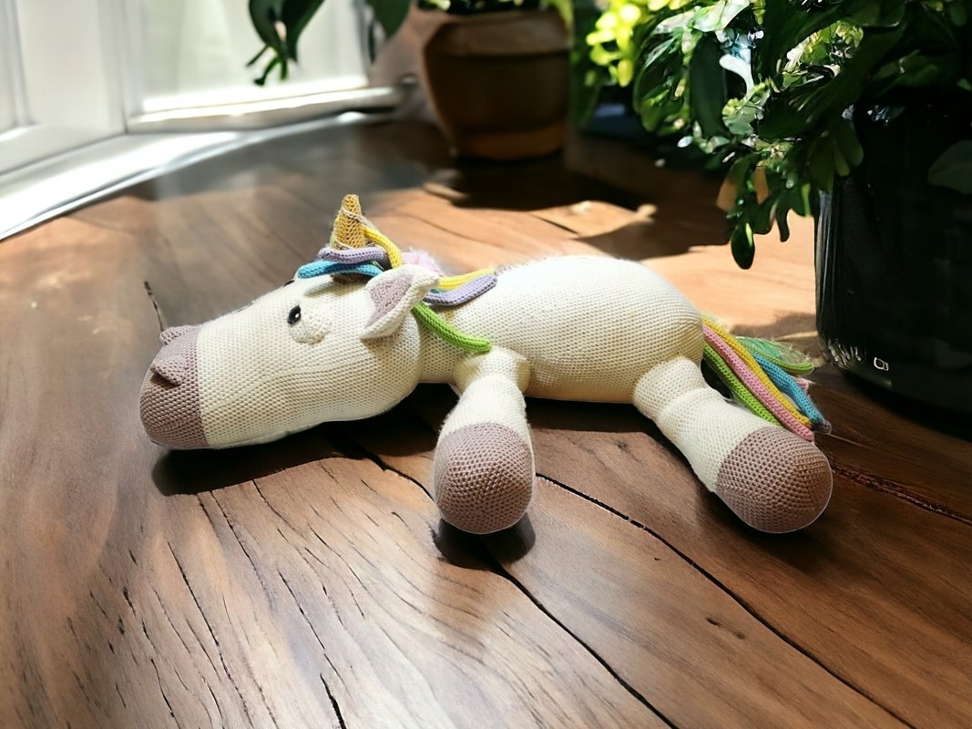 Cream crocheted amigurumi unicorn with pastel rainbow mane and tail on a wooden surface.