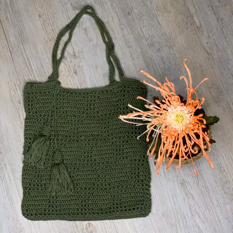 Olivgrüner gehäkelter Shopper mit langen Henkeln und zwei Quasten neben einem Korb mit orangefarbener Blüte auf hellem Holz.