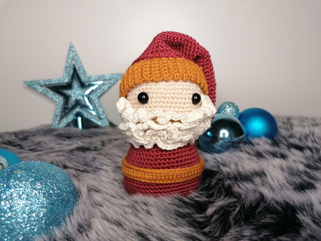 Gehäkelter Amigurumi-Weihnachtsmann mit roter Mütze und weißem Bart auf grauem Kunstfell, umgeben von blauen Weihnachtskugeln und Stern