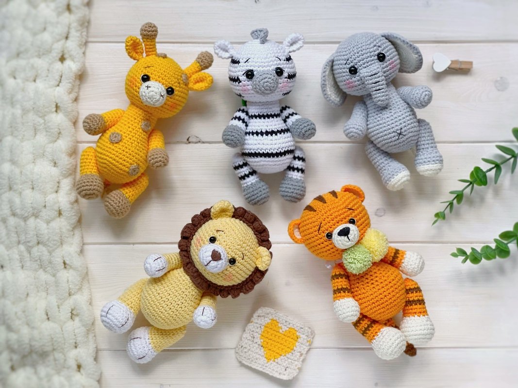 Flat-lay of five crocheted amigurumi animals: yellow giraffe, striped zebra, grey elephant, lion with mane and orange tiger on a light wood background