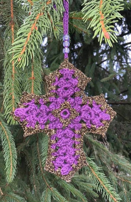Gehäkeltes lila-goldenes Kreuz an einem Tannenzweig, mit sichtbaren Häkelstrukturen und Perlenaufhängung.