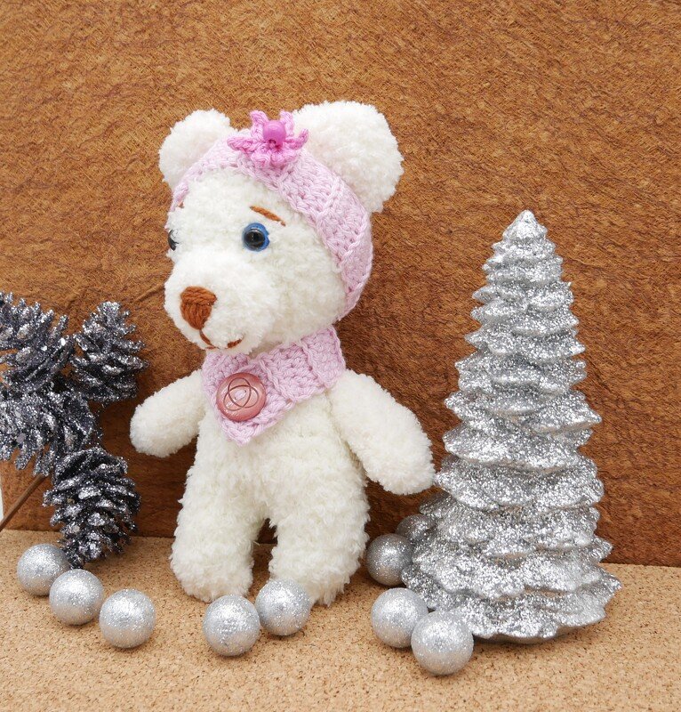 Weißer Amigurumi-Teddy mit rosa Häkelmütze und Kragen, umgeben von silbernen Kugeln, Schneezapfen und einem Mini-Weihnachtsbaum