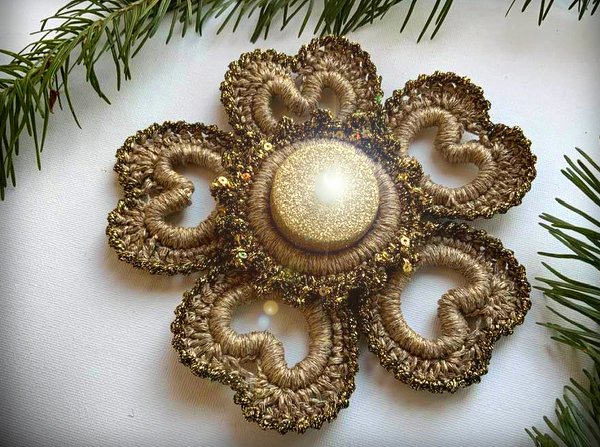 Gehäkelte goldbraune Deko-Blüte mit glitzerndem Zentrum und Pailletten vor hellem Hintergrund und Tannenzweigen