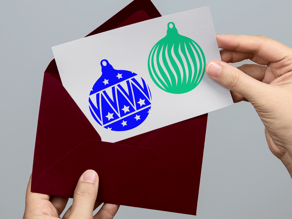 Karte mit zwei Plottmotiven: blaues Weihnachtskugel-Design mit Sternen und Zickzack und grünes Kugel-Design mit geschwungenen Streifen, Hand hält Karte über dunkelrotem Umschlag.