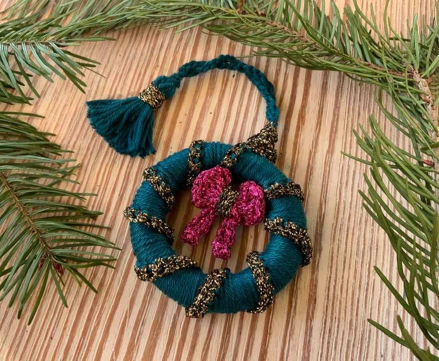 Mini-Häkelkranz in Petrol mit goldener Borte, pinker Schleife und Quaste auf Holz, umgeben von Tannenzweigen