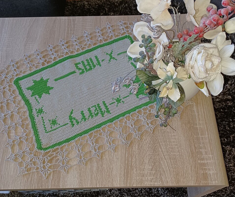Gehäkelter Tischläufer creme/hellgrau mit grünem 'Merry X‑Mas' Schriftzug, Sternen und Spitzenrand auf hellem Holztisch neben einer Blumen­dekoration.