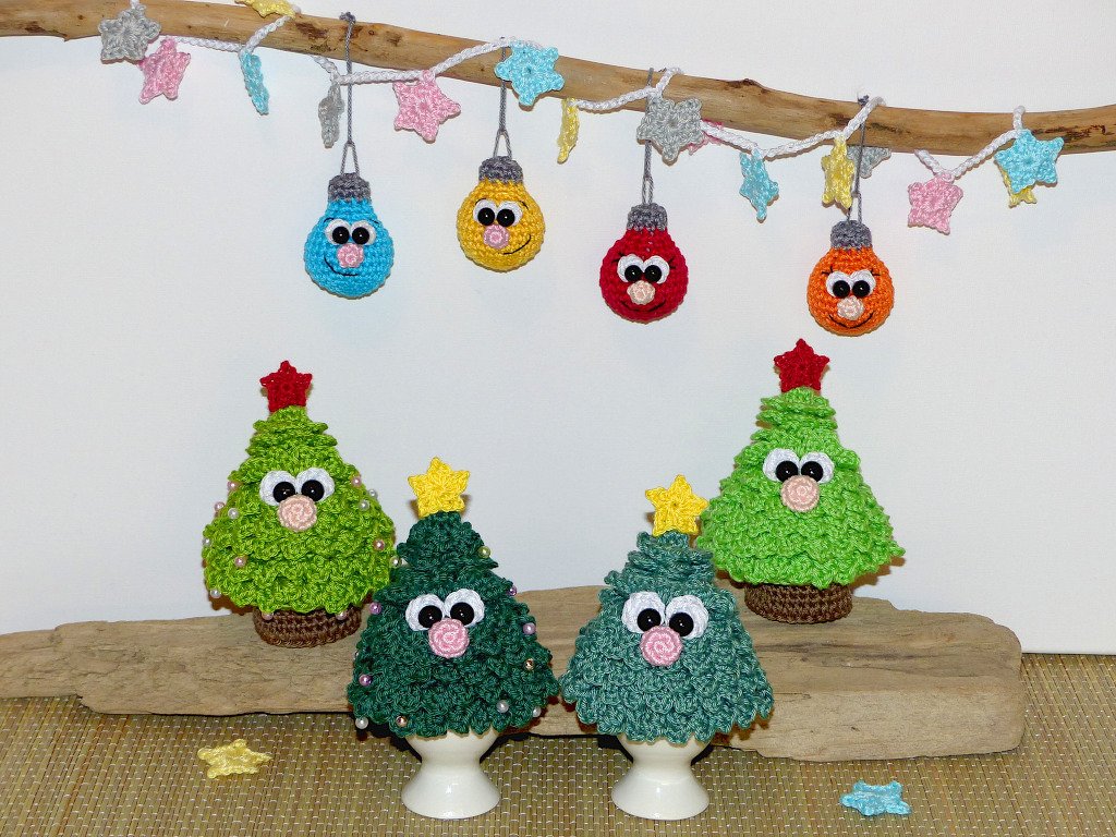 Mehrere gehäkelte Mini-Weihnachtsbäume mit Perlen und Sternspitzen, zwei in Eierbechern, darüber Girlande mit bunten Häkelkugeln an einem Ast