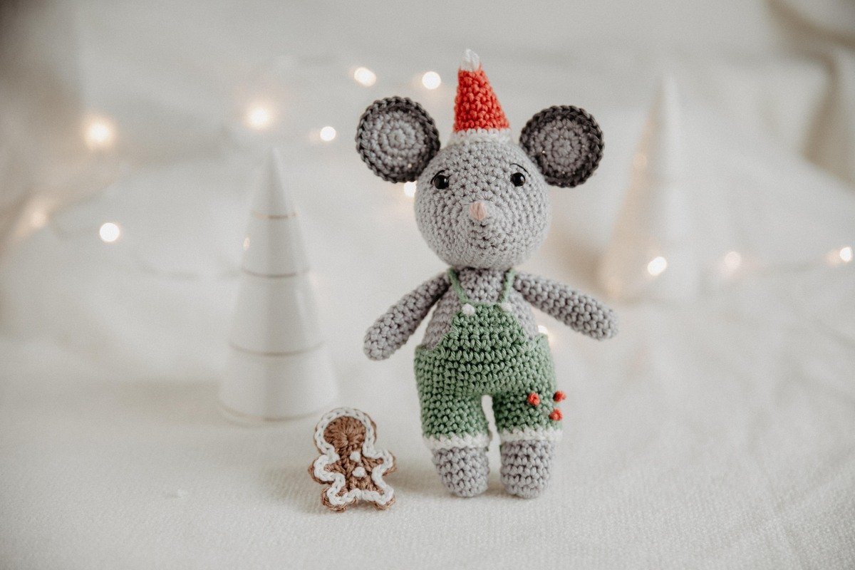 Grau gehäkelte Amigurumi‑Maus mit roter Mütze und grüner Latzhose neben kleinem gehäkelten Lebkuchenmann vor hellem Stoff und Lichterbokeh.