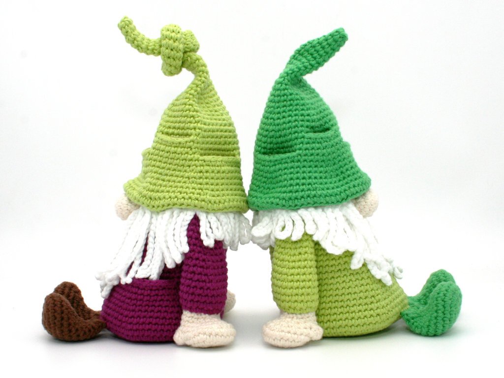 Two crocheted gnomes seen from behind wearing different green hats and white beards, seated on a white background.