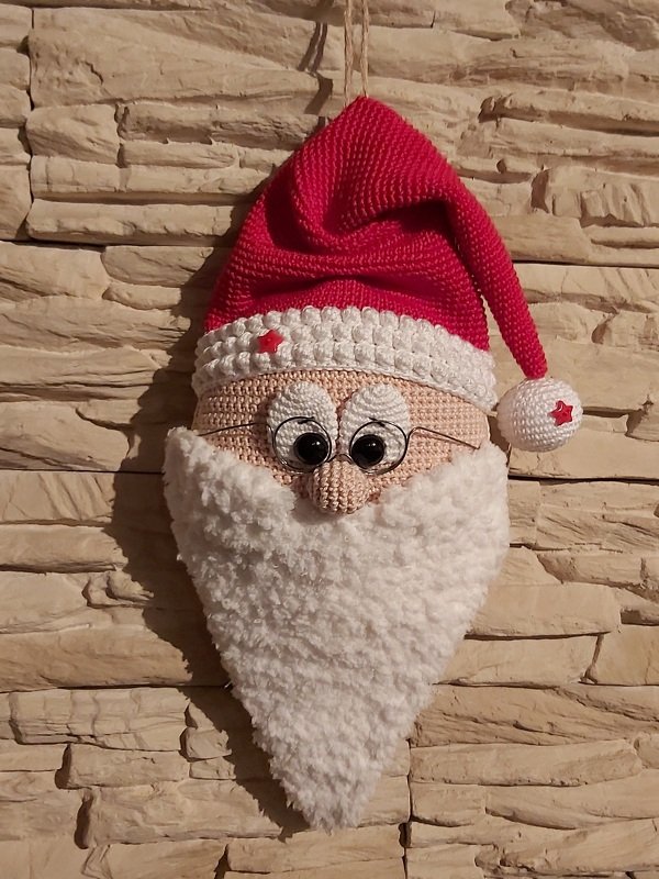 Gehäkelter Weihnachtsmannkopf mit roter Mütze und weißem Bart an Steinwand