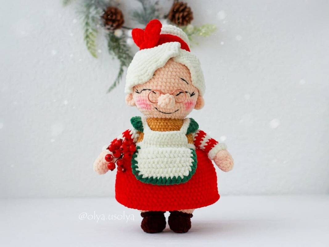 Gehäkelte Amigurumi-Weihnachtsfrau mit Brille, weißem Haar, rotem Kleid, weißer Schürze und roten Beeren