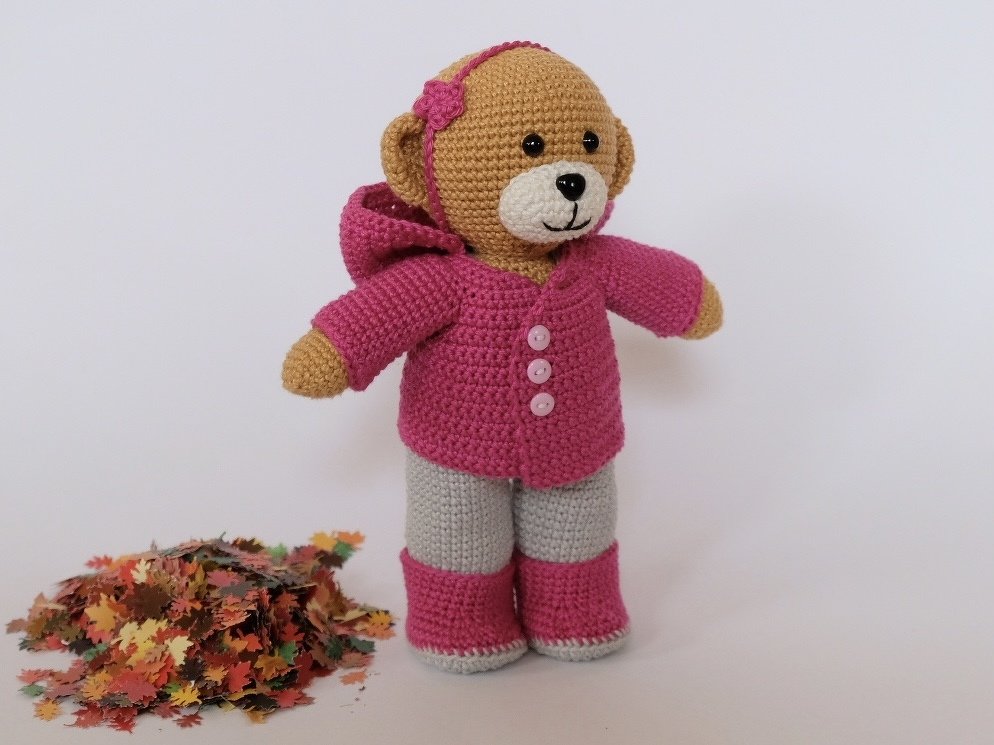 Gehäkelter Amigurumi-Bär mit Knöpfen an der Jacke und zweifarbigen Hosen neben Herbstblättern