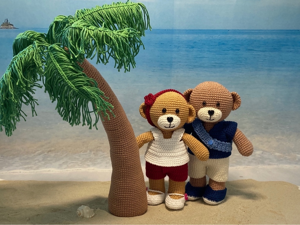 Zwei gehäkelte Amigurumi-Teddys in Sommeroutfits neben gehäkelter Palme vor Strandkulisse