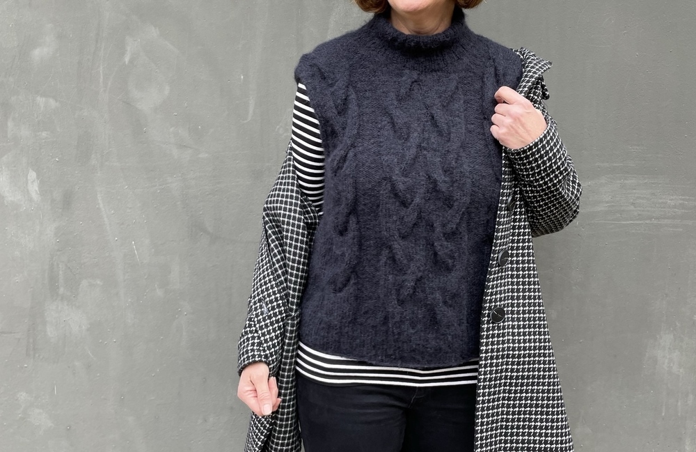 Ärmellose dunkelgraue Strickweste mit Zopfmuster und Stehkragen, getragen über gestreiftem Langarmshirt; kariertes Mantelteil über der Schulter.
