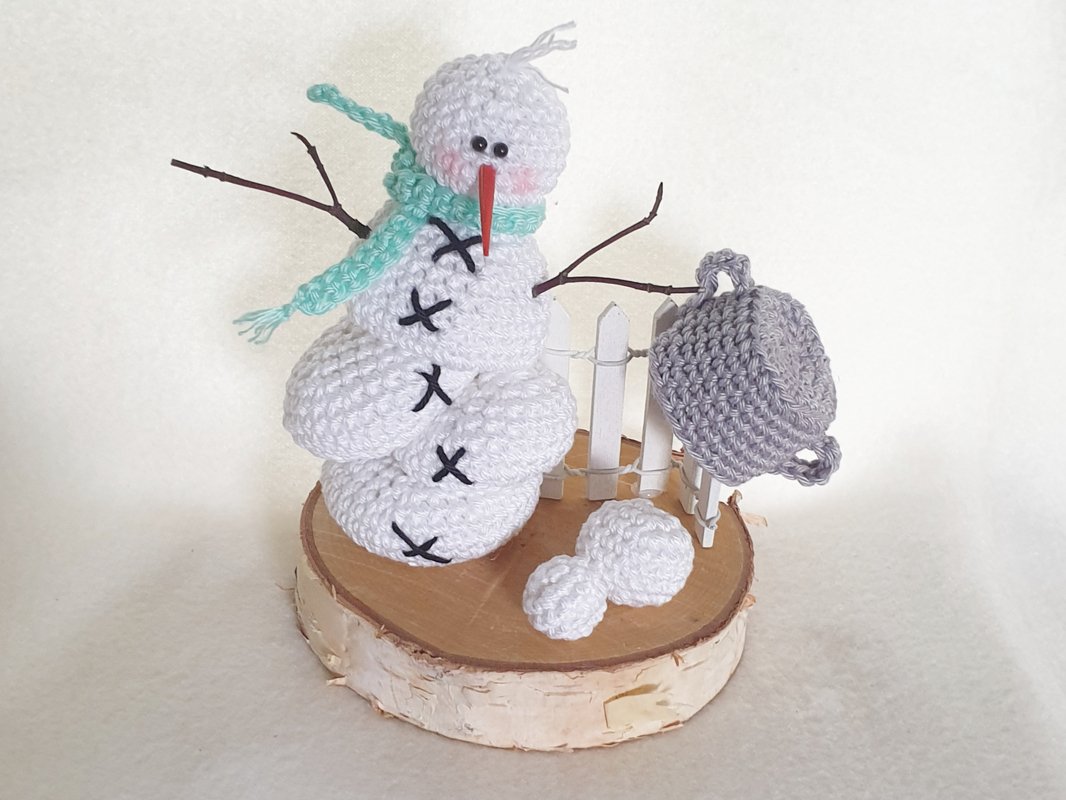 Gehäkelter weißer Amigurumi‑Schneemann mit türkisfarbenem Schal auf Birkenholzscheibe
