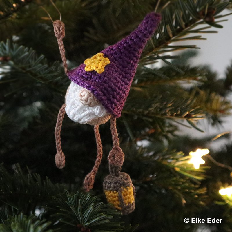 Crocheted amigurumi gnome ornament with purple hat and small lantern hanging on a fir branch