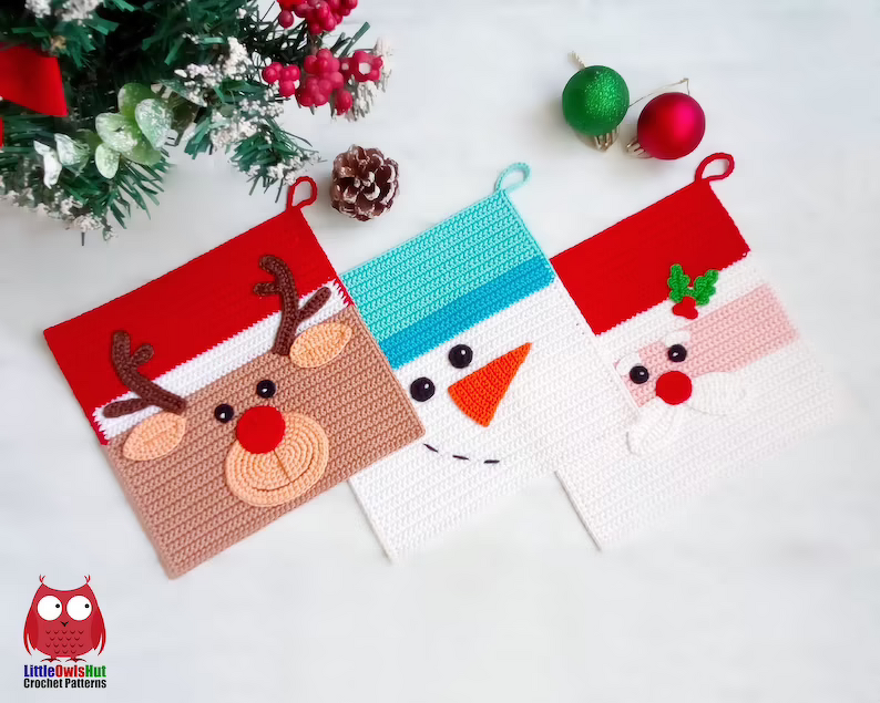 Drei gehäkelte Weihnachts-Topflappen mit Rentier-, Schneemann- und Weihnachtsmann-Motiv, arrangiert mit Tannengrün und Christbaumkugeln.