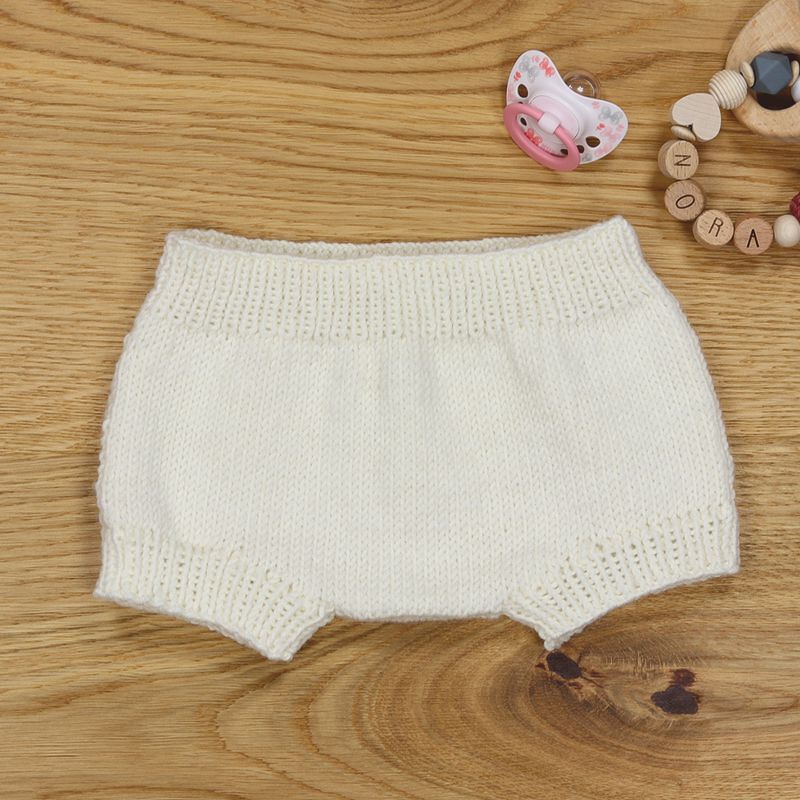 Cremefarbene gestrickte Babyhose auf Holzuntergrund neben Schnuller und Greifling