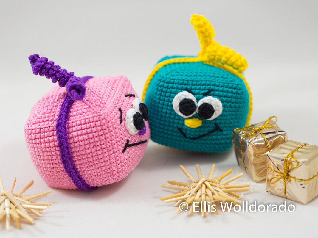 Zwei gehäkelte Geschenkfiguren in Pink und Türkis mit aufgenähten Augen, daneben goldene Päckchen und Strohsterne auf hellem Hintergrund.
