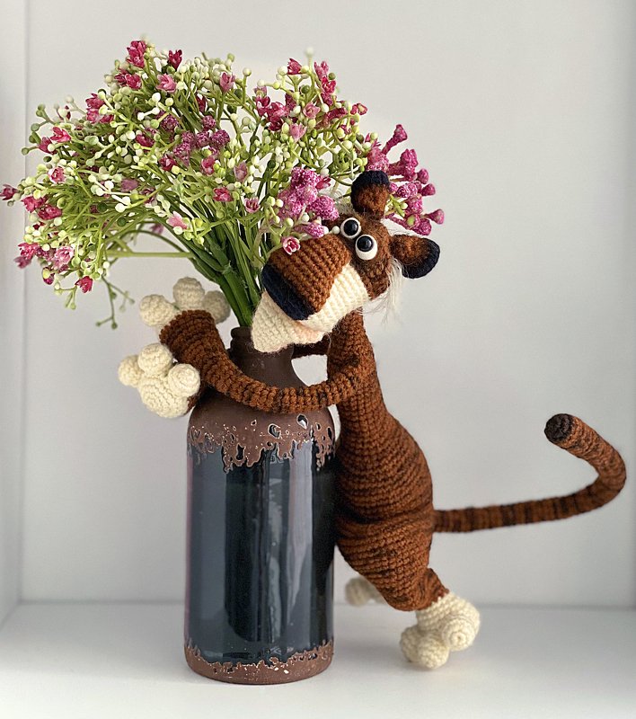 Nahaufnahme eines braunen gehäkelten Amigurumi mit langen Armen und gestreiftem Schwanz neben einer Vase