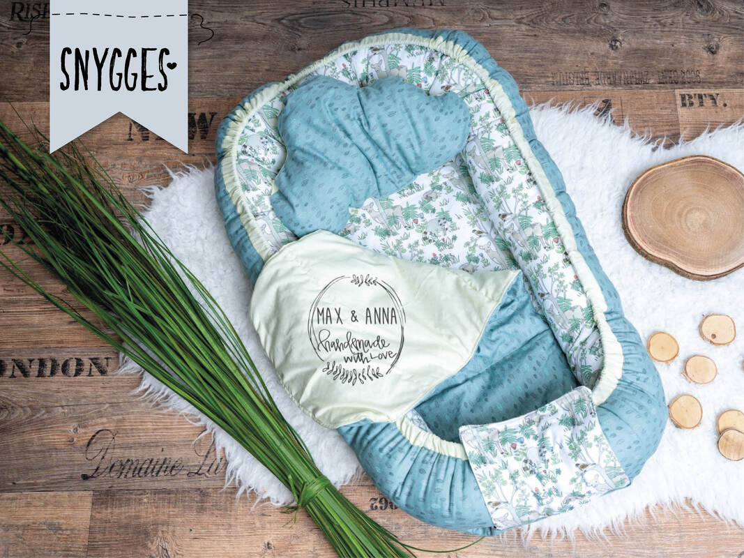 Blau-grünes Babynest mit floralem Innenfutter, Wolkenkissen und Einschlagdecke mit Schriftzug auf Holzfußboden