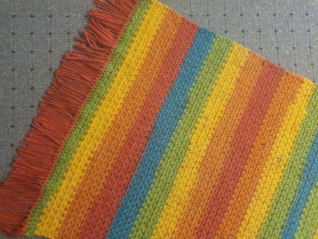 Gehäkelte, gestreifte Decke in Regenbogenfarben mit Fransen auf grauem Untergrund