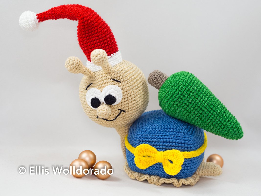 Gehäkelte Amigurumi‑Schnecke mit roter Weihnachtsmütze, blauem Schneckenhaus, gelber Schleife und grünem Tannenbaum