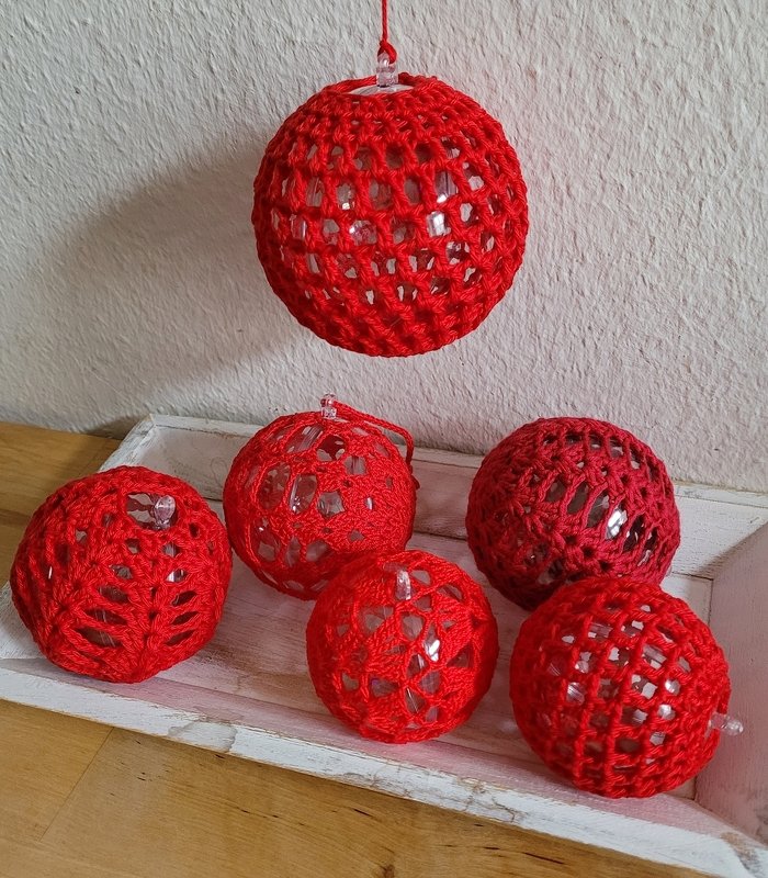 Sechs rote gehäkelte Kugeln mit transparenten Perlen in einem Netzmuster. Eine Kugel hängt, die anderen liegen in einer weißen Schale.