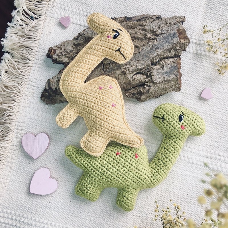 Two amigurumi dinosaur dolls, one beige and one green, on a textured white blanket with small pink hearts and a piece of wood.