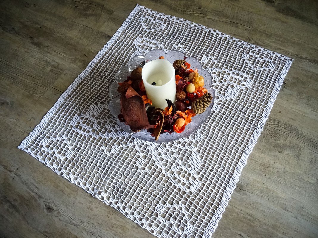 Weißer gehäkelter Tischläufer mit durchbrochenem Filetmuster und einer Dekoschale mit Kerze und Potpourri auf einer Holzfläche.