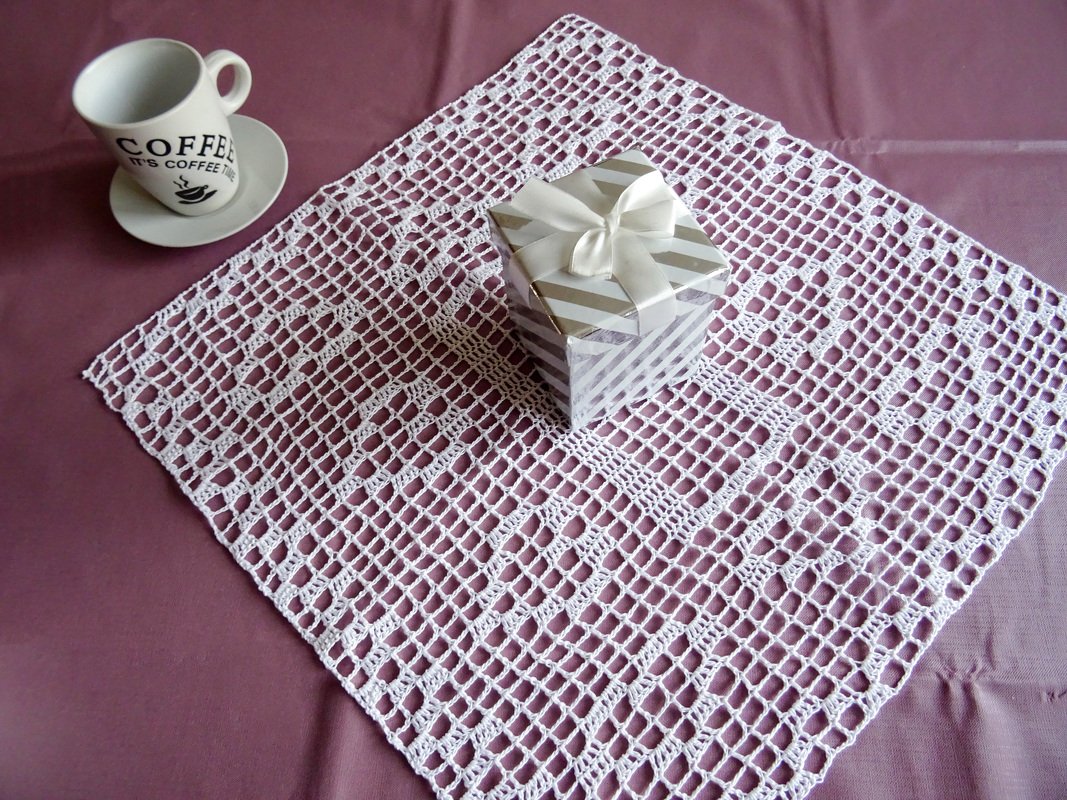 Weißes rechteckiges Häkeldeckchen im Filetgitter auf lila Stoff, daneben Kaffeetasse und Geschenk.