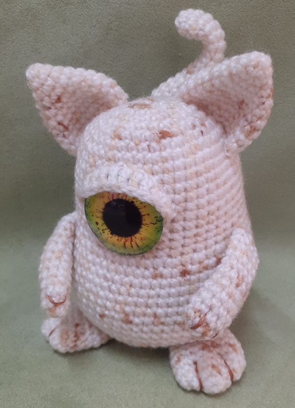 Cream-speckled crocheted amigurumi monster with one large green-and-yellow eye, pointy ears and a curled tail on a pale green background.