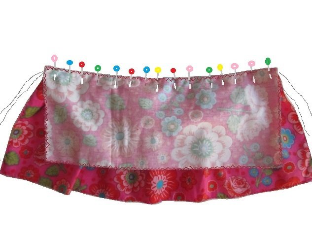Zwei geblümte Stofflagen, eine hellrosa und eine dunkleres Rosa, mit bunten Stecknadeln am oberen Rand geheftet und mit Zickzack- oder Overlockstich versäubert.