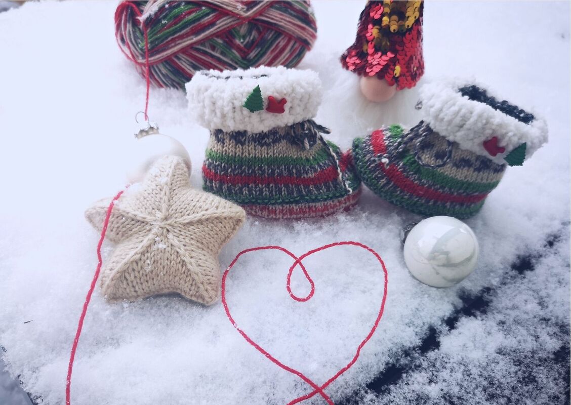Gestrickte Babyschuhe mit grün-roten Streifen und weißem, flauschigem Umschlag auf Schnee, gestrickter Stern und rotes Garnherz