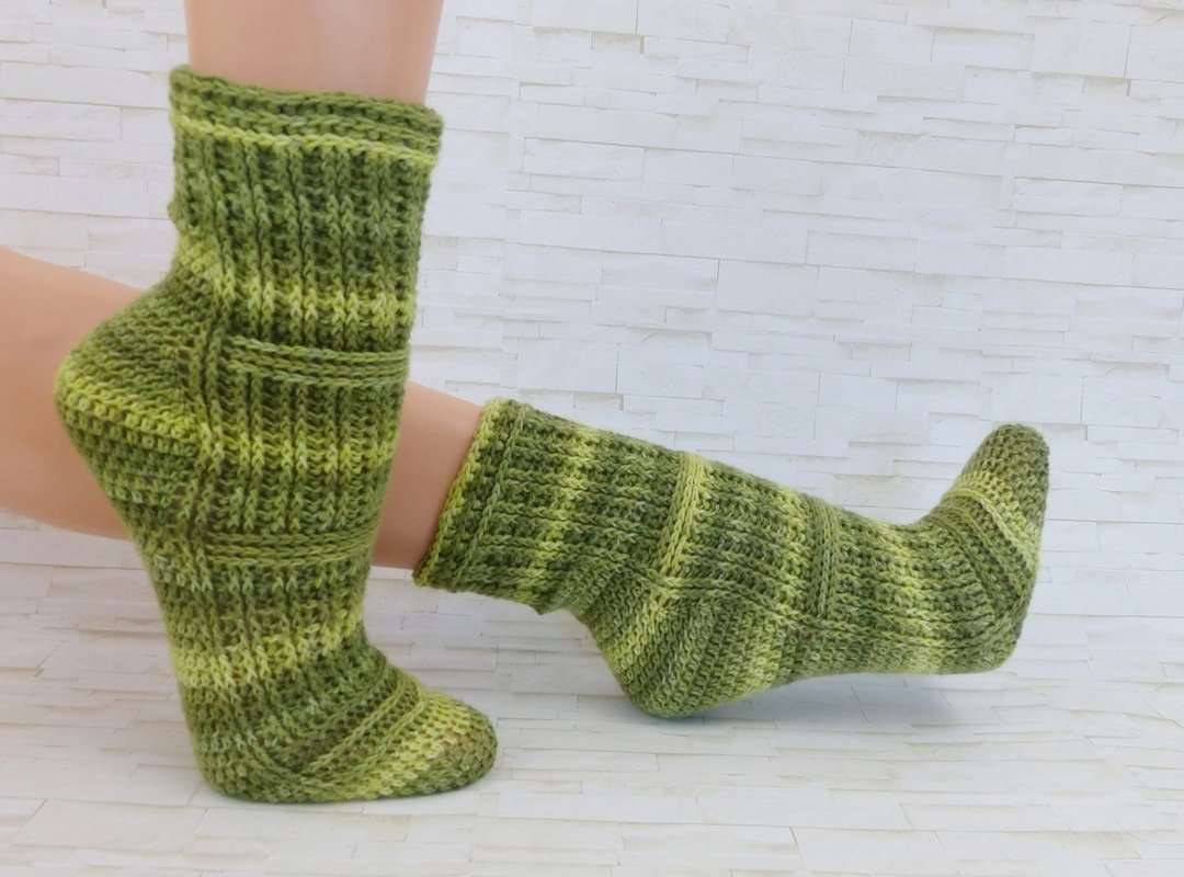 Zwei handgehäkelte grüne Socken mit Rippen- und Strukturmuster an ausgestellten Füßen vor hellem Hintergrund.