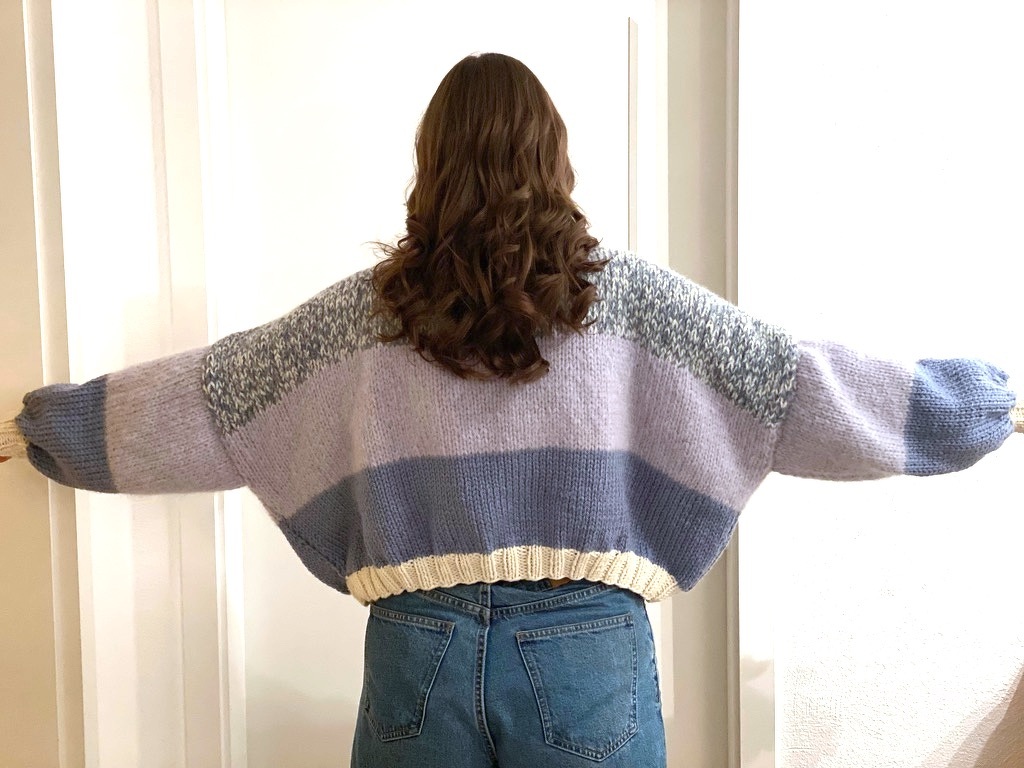 Rückenansicht einer Person in kurz geschnittener Oversize-Strickjacke mit pastellfarbenen Blockstreifen und ausgestreckten Armen