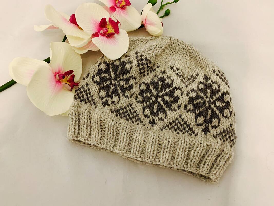 Beige gestrickte Damenmütze mit dunkelgrauem Sternmuster und Rippbündchen neben weißen Orchideen