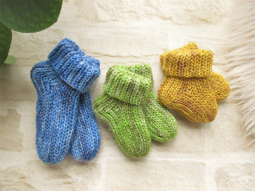 Drei handgehäkelte Babysocken in Blau, Grün und Gelb mit umgeschlagenem Bündchen auf hellem Untergrund